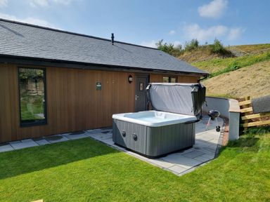 Outdoor patio area with a hot tub and a barbecue next to a wooden house at Meadow View in Meifod Powys