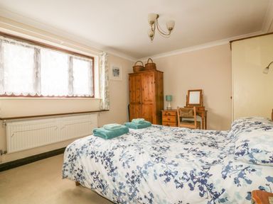 A bedroom with a bed covered in floral bedding towels on the bed a wooden wardrobe and a dressing table with a chair and lamp