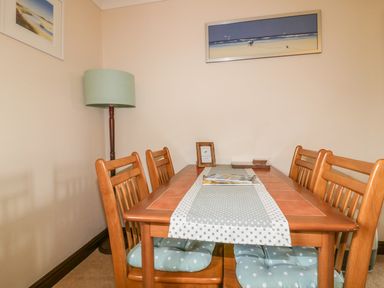 A dining area with a wooden table and six chairs with cushions a table runner a floor lamp and two framed pictures on the walls at Chiffchaff in Padstow