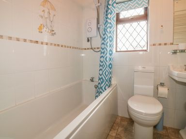 A bathroom with a bathtub shower toilet sink and a window with a blue patterned curtain at Chiffchaff in Padstow