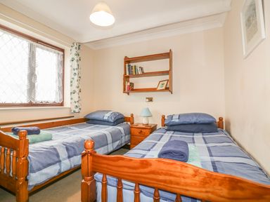 A bedroom with two single beds a wooden nightstand with a lamp a wooden wall shelf with books and a window with curtains at Chiffchaff in Padstow