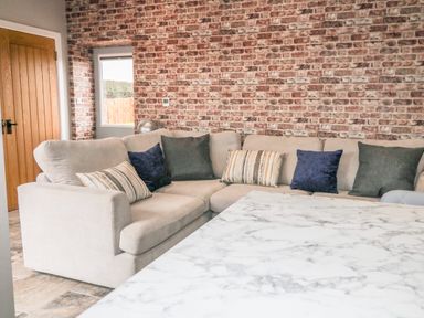 A living room with a beige sectional sofa with cushions a brick wall a wooden door and a marble table at Owl View in Lebberston near Cayton