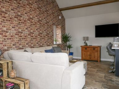 A living room with a beige sofa set against a brick wall a wooden cabinet with a lamp and a mounted black TV at Owl View in Lebberston near Cayton