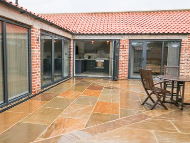 An outdoor patio area with stone tiles a round wooden table and chairs and a view into a kitchen and living space through glass doors at Owl View in Lebberston near Cayton