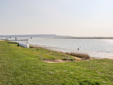 A grassy shoreline with a small boat on the shore a wooden pier and boats in the water at Harbour View Mudeford in Christchurch