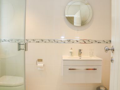 A bathroom with a wall-mounted sink and cabinet a round mirror a toilet paper holder and a glass shower door at Harbour View Mudeford in Christchurch