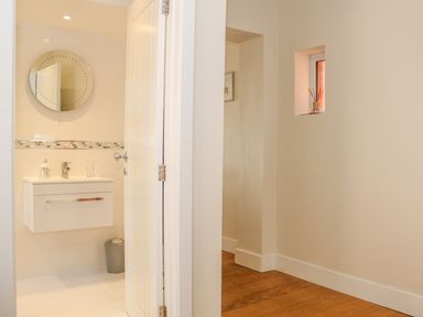 A bathroom with a round mirror and white sink visible through an open door in a hallway with wooden flooring and a small window at Harbour View Mudeford in Christchurch