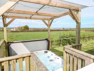 An outdoor hot tub under a wooden pergola with string lights and a view of fields at Mountain View Lodge Tavernspite near Narberth