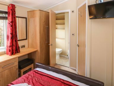 A bedroom with a wooden wardrobe, desk with a stool, red curtains, a bed with a red bedspread, and a door leading to a bathroom at Mountain View Lodge in Tavernspite near Narberth