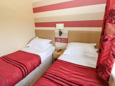 A bedroom with two single beds with red bedspreads a bedside table with a remote control a wall lamp and striped wallpaper at Mountain View Lodge Tavernspite near Narberth