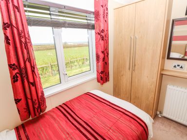 A bedroom with a bed covered in red and white bedding next to a window with red curtains and a wooden wardrobe at Mountain View Lodge Tavernspite near Narberth