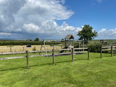 A playground with swings and slide behind a wooden fence in a grassy area with fields and a tree in the background at Mountain View Lodge in Tavernspite near Narberth