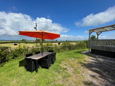 A garden area with a table and chairs under an orange umbrella near a wooden deck with farmland in the background at Mountain View Lodge in Tavernspite near Narberth