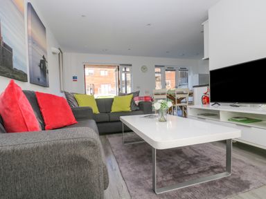 A living room with gray sofas and colorful cushions a white coffee table a television and dining table at Sea Breeze 17 Gannel in Porthtowan