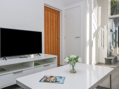 A TV on a white stand near two doors with a white table and flowers and a magazine with stairs and a window in the background at Sea Breeze 17 Gannel in Porthtowan