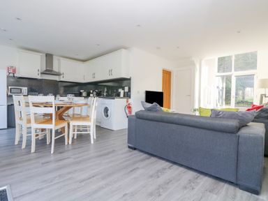 An open plan room with a kitchen area featuring white cabinets a dining table with chairs and a living area with a grey sofa at Sea Breeze 17 Gannel in Porthtowan