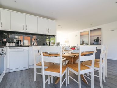 A kitchen with white cabinets a dishwasher and a wooden dining table with white chairs at Sea Breeze 17 Gannel in Porthtowan