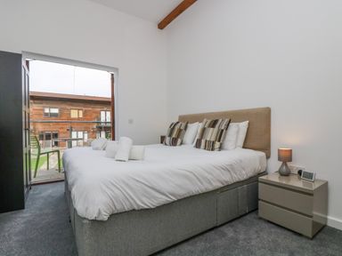 A bedroom with a double bed pillows bedside table lamp alarm clock and open door to balcony at Sea Breeze 17 Gannel in Porthtowan