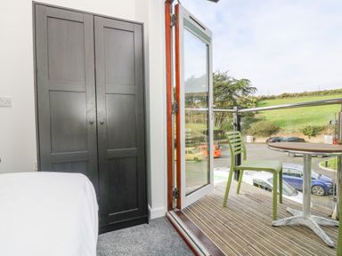 A bedroom with a dark wardrobe next to an open door leading to a balcony with a green chair and round table at Sea Breeze 17 Gannel in Porthtowan
