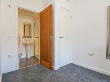 A room with carpeted floor showing a wooden door open to a bathroom with a sink and shower and a closed white door at Sea Breeze 17 Gannel in Porthtowan