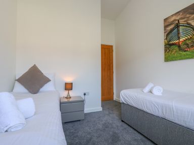 A bedroom with two single beds, a bedside table with a lamp, and a painting of a boat at Sea Breeze 17 Gannel in Porthtowan
