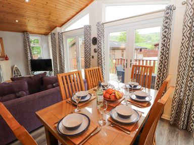 A dining table set with plates and glasses next to a living area with purple sofas and large windows with patterned curtains at Fell View Lodge in Troutbeck