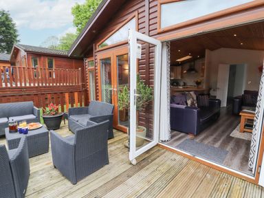 An outdoor wooden deck with black woven patio furniture and a potted plant attached to a wooden house with open glass doors revealing a living room with dark sofas at Fell View Lodge in Troutbeck