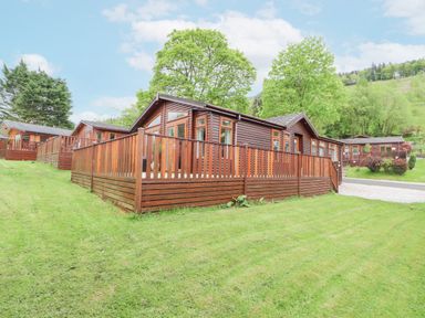 A wooden lodge with a fenced deck surrounded by grass and trees at Fell View Lodge in Troutbeck