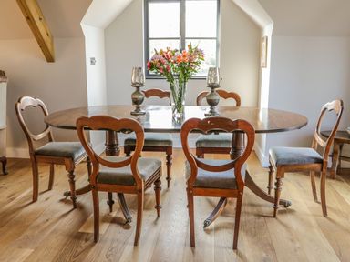 A dining room with a wooden table and six wooden chairs with a vase of flowers and two candle holders on the table at The Coach House in St Columb Major