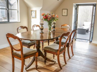 A dining room with a wooden table and six chairs with a vase of flowers on the table and doors leading to an outdoor seating area at The Coach House in St Columb Major