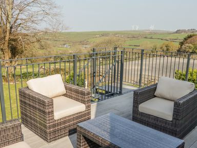 A patio with two cushioned chairs and a table overlooking a rural landscape with hills and wind turbines at The Coach House in St Columb Major