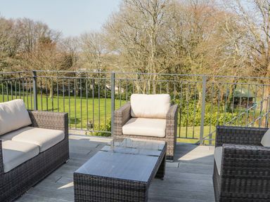 An outdoor seating area with wicker furniture and a glass top table on a balcony overlooking a grassy area with trees at The Coach House in St Columb Major