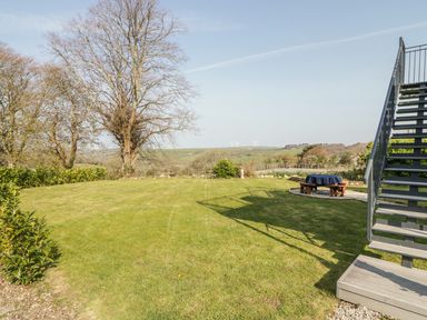 A lawn with trees and a picnic table covered with a cloth near metal stairs in a rural area with wind turbines in the distance at The Coach House in St Columb Major
