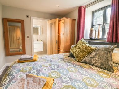 A bedroom with a patterned bedspread pillows towels a wooden wardrobe a wall mirror and an open door leading to a bathroom at The Coach House in St Columb Major