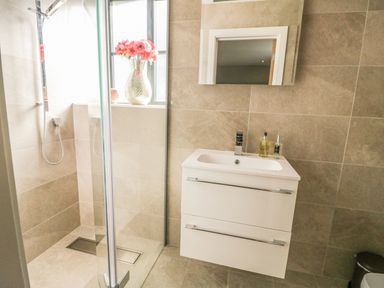 A bathroom with a glass shower enclosure a white sink cabinet with a mirror above and a vase with flowers on the window sill at The Coach House in St Columb Major