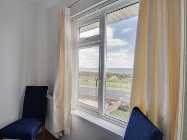 A window with cream curtains and two blue chairs facing it showing a view of the sea at The Lookout -The House With A View in Weymouth