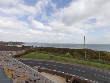 A view of a road with a grassy area and benches overlooking the sea with houses and bushes in the background at The Lookout -The House With A View in Weymouth