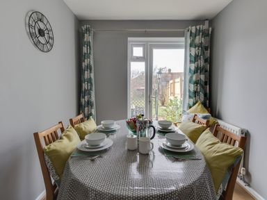 A dining room with a set table and wooden chairs near glass doors with patterned curtains at The Lookout -The House With A View in Weymouth