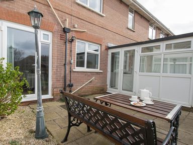 A patio area with a wooden bench and table set with teapot and cups outside a brick house with conservatory at The Lookout -The House With A View in Weymouth