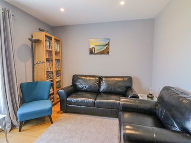 A living room with black leather sofas a blue armchair a wooden bookshelf and a wall painting at The Lookout -The House With A View in Weymouth
