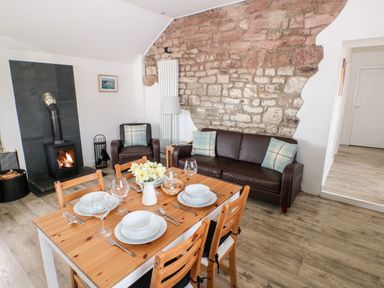 A dining area with a wooden table set with plates and glasses next to a sitting area with a leather sofa and armchair beside an exposed stone wall and a wood burning stove at Dairy Cottage Tavernspite near Narberth