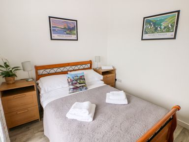 A bedroom with a bed with a gray bedspread and folded towels two wooden bedside tables with lamps and a plant and two framed pictures on the walls at Dairy Cottage Tavernspite near Narberth