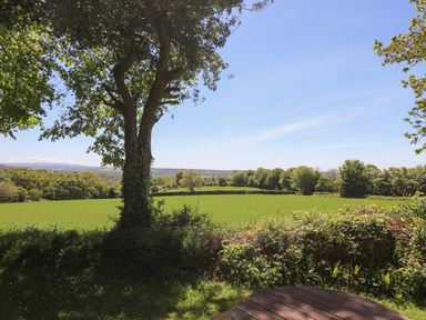 A view of a green field with trees and a blue sky from a garden with a wooden table at Dartmoor 2 in Callington