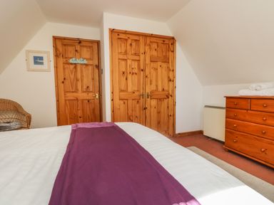 A bedroom with a bed covered with white and purple linens a wooden dresser with towels on top wicker chair wooden doors and white walls at Dartmoor 2 in Callington