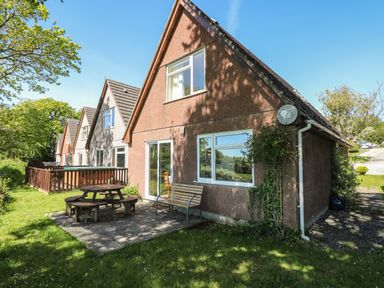A row of houses with triangular roofs a wooden picnic table and bench on a paved patio with grass and trees at Dartmoor 2 in Callington