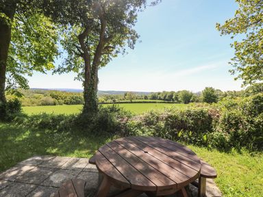 A wooden round picnic table on a stone patio overlooking green fields and trees at Dartmoor 2 in Callington