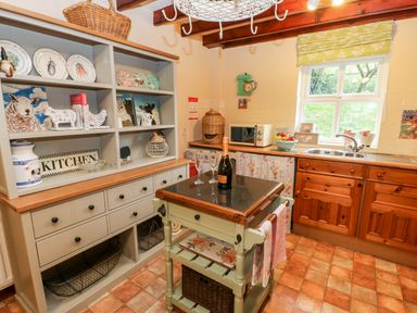 A kitchen with wooden cabinets a tiled floor a small island with a bottle and two glasses a shelf unit with decorative plates and items at Isaacs Cottage Spartylea near Allendale
