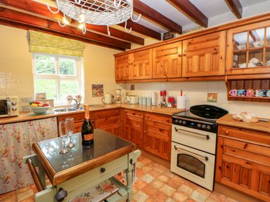 A kitchen with wooden cabinets a stove a small table with a bottle and glasses and a window with a floral curtain at Isaacs Cottage Spartylea near Allendale