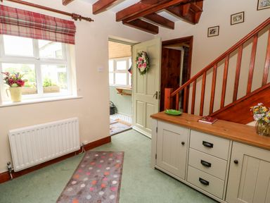 An entrance hallway with a wooden staircase a cabinet with drawers and flowers on top a window with a floral vase and a doorway leading to another room at Isaacs Cottage Spartylea near Allendale