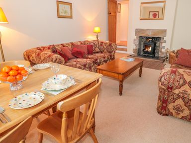 A living room with patterned sofas a wooden coffee table a dining table set with plates bowls and a bowl of oranges and a lit fireplace at West Burnbank in Tarset near Bellingham